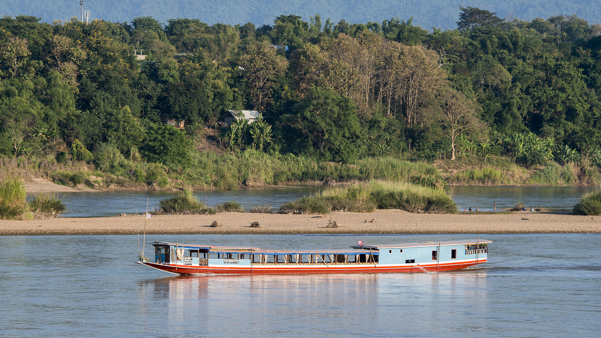 Long wooden slow boat on the Mekong beneath forested hills