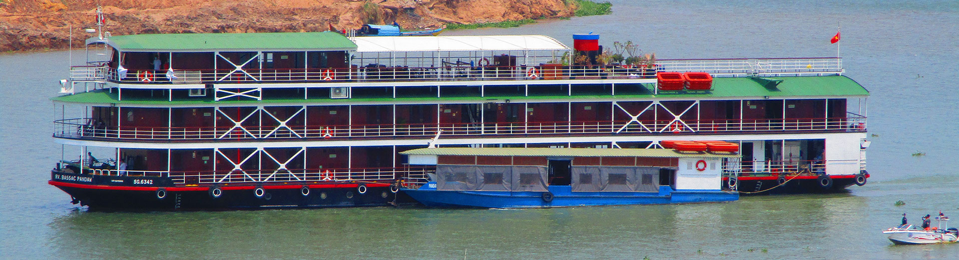 Multi-deck river cruise boat with a red hull and green roof on the river